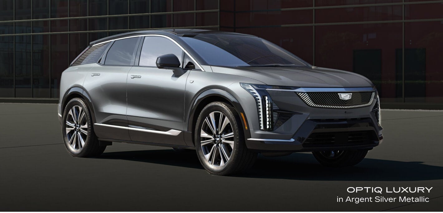 Front-three-quarter view of a Cadillac OPTIQ Luxury in Argent Silver Metallic parked in front of a modern glass building.